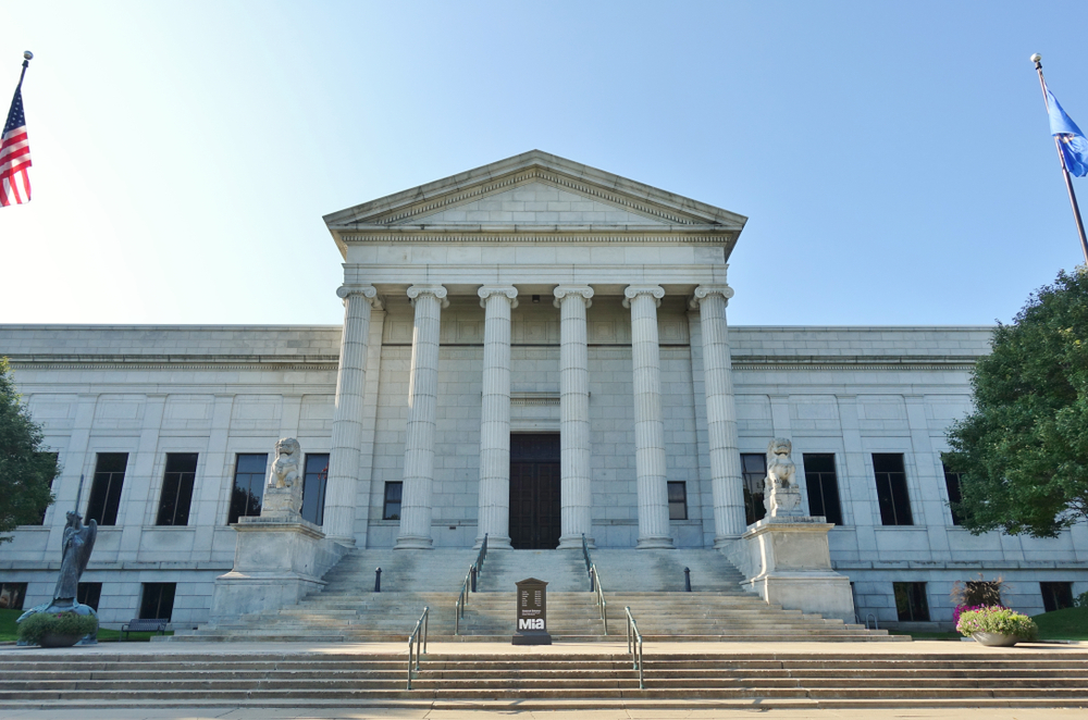 The front entrance to the Minneapolis Institute of Art. It is a large Roman style building made of stone with tall columns on the front. It has steps leading to the front door and a pair of carved stone lions on either side. 