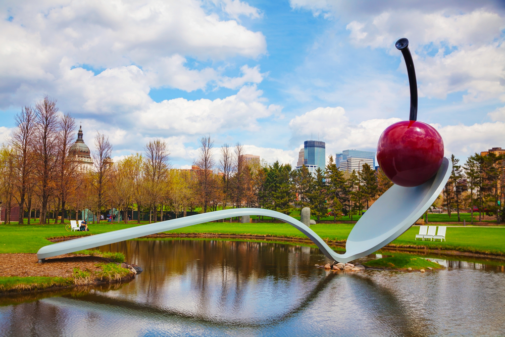 A large sculpture of a spoon with a cherry perched on top of it. The spoon is resting on the shore and crosses to the middle of a small lake. Around it is a grassy area with benches and trees in the distance with little to no leaves. Behind the trees you can see a city skyline. One of the best things to do in Minnesota. 