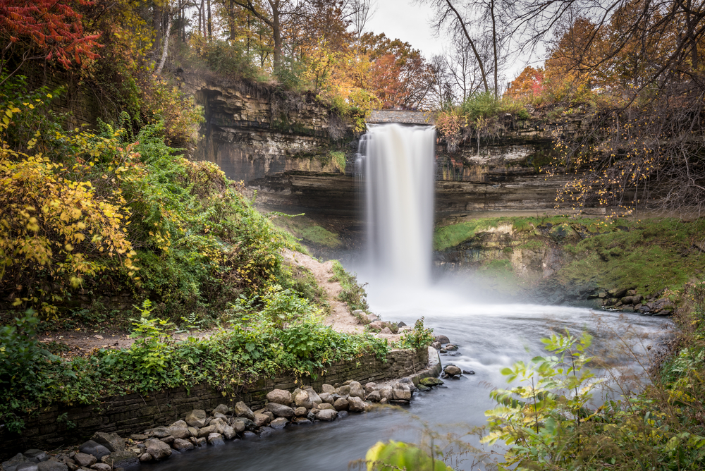 A bridal veil fall that is flowing into a river. it is surrounded by rocky cliffs, grasses, and trees. There is moss growing on the rock and the trees have red, yellow, green, and red leaves. One of the best things to do in Minnesota.
