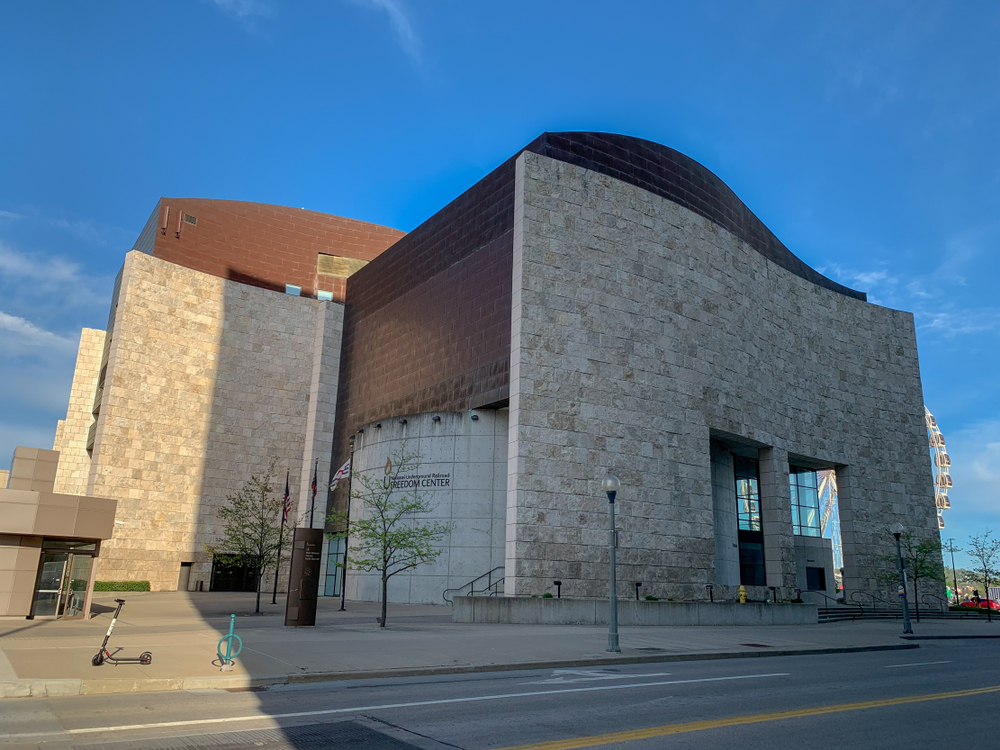 The exterior of the National Underground Railroad Freedom center. It is a stone building with different angles and a dark brick roof and siding. Behind it you can see a small ferris wheel and in front of it is a courtyard with small trees. 