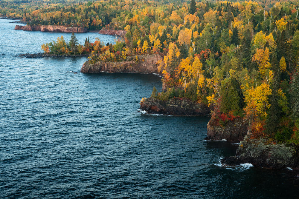 A rocky shore line along Lake Superior, one of the best things to do in Minnesota. On the rocky cliffs there are trees growing. They have red, yellow, orange, and green leaves. 