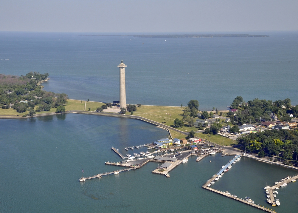 An aerial view of Put-in-Bay on South Bass Island. You can see a large stone tower memorial, grassy areas with trees, a few buildings, and a boat marina. The island is surrounded by water in Lake Erie. 