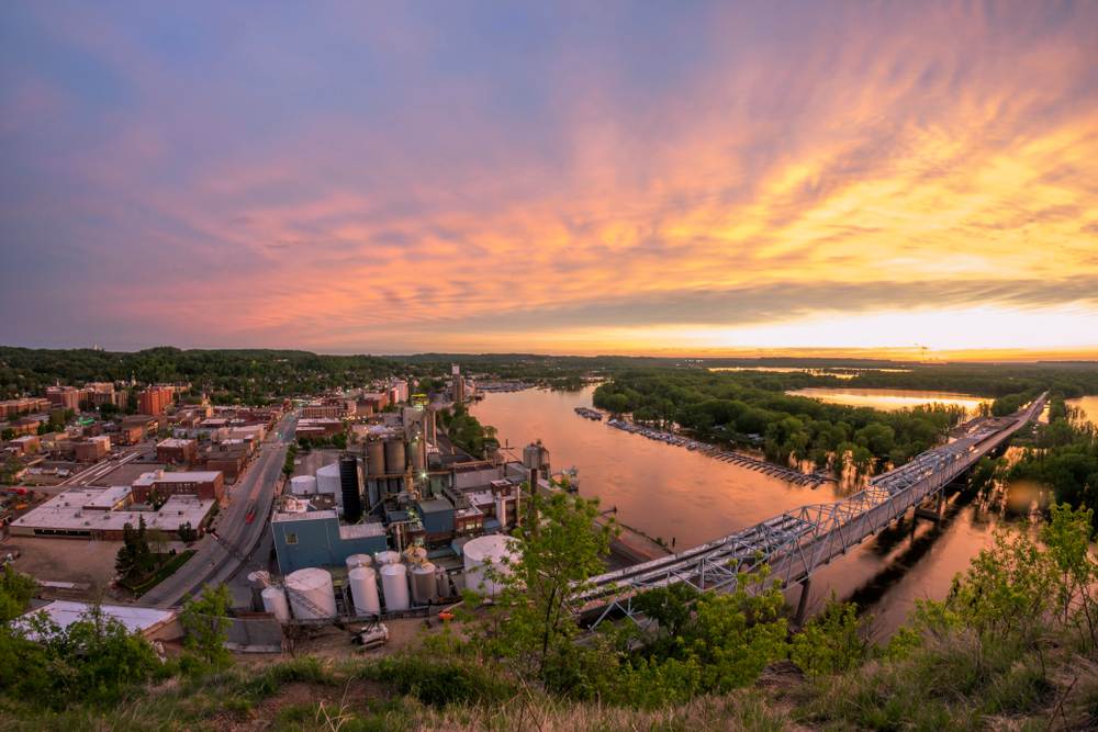 An aerial view of Downtown Red Wing. You can see a bridge crossing the river, factories, and other buildings. In the distance there are trees and there are trees along the river. It is sunset and the sky is orange, yellow, pink, and blue. 