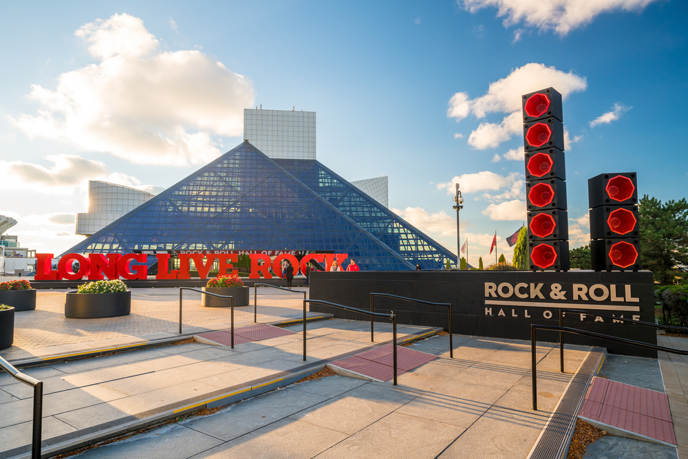 The exterior of the Rock and Roll Hall of Fame, one of the best Ohio day trips. It is a pyramid shaped glass building with a big red sign that says 'long live rock' in front of it. You can also see a sign that says 'Rock and Roll Hall of Fame' and looks like it has black and red speakers on it. 