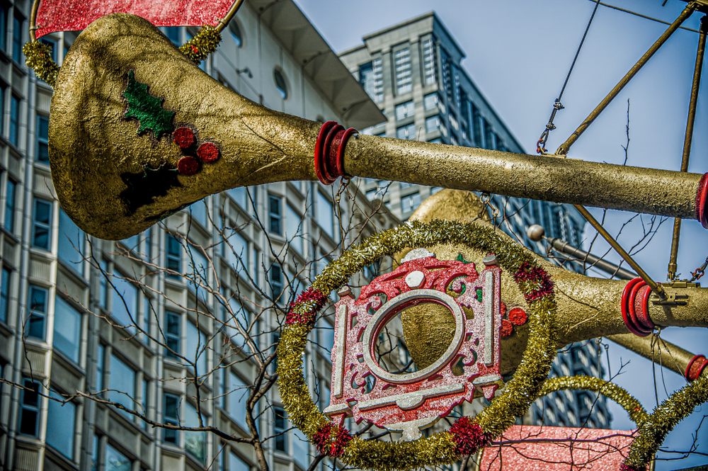 Gold trumpets with holly and other holiday decorations on them hanging from a street light. Behind them you can see a skyscraper. 