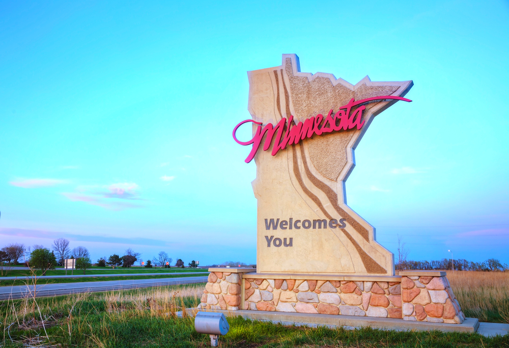 A sign on the side of a highway welcoming you to Minnesota. It is a large stone in the shape of the state and Minnesota is in red cursive.