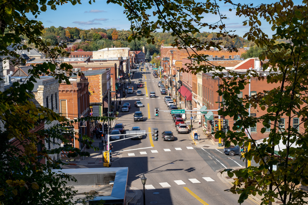 A slight aerial view of Downtown Stillwater. It is a charming main street with brick buildings, cars parked on the side of the road, and trees around the buildings. 