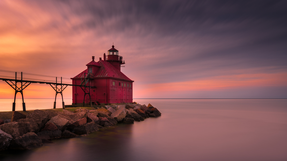 A red lighthouse on the end of a stone pier in the middle of a lake. There are power lines leading to the lighthouse. The water surface is calm and the sun is setting so the sky is orange, yellow, pink, and purple. 