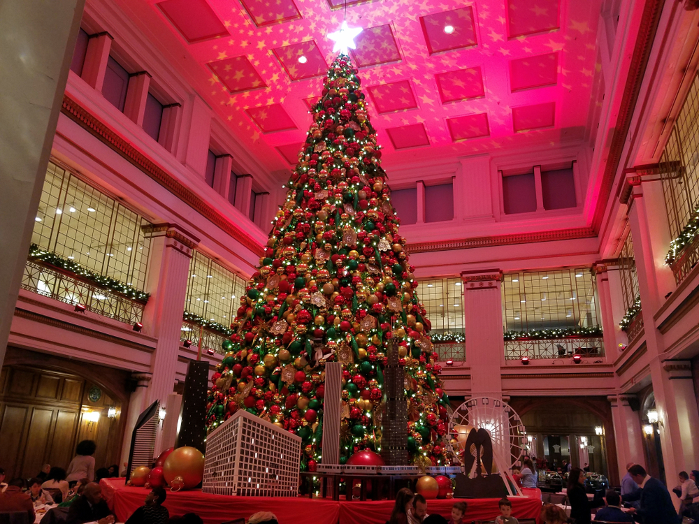 A large elaborately decorated Christmas tree on a pedestal in the middle of a large room. The room is colored with redish pink lights, there are windows high on the wall, and people are sitting at tables around the tree. 