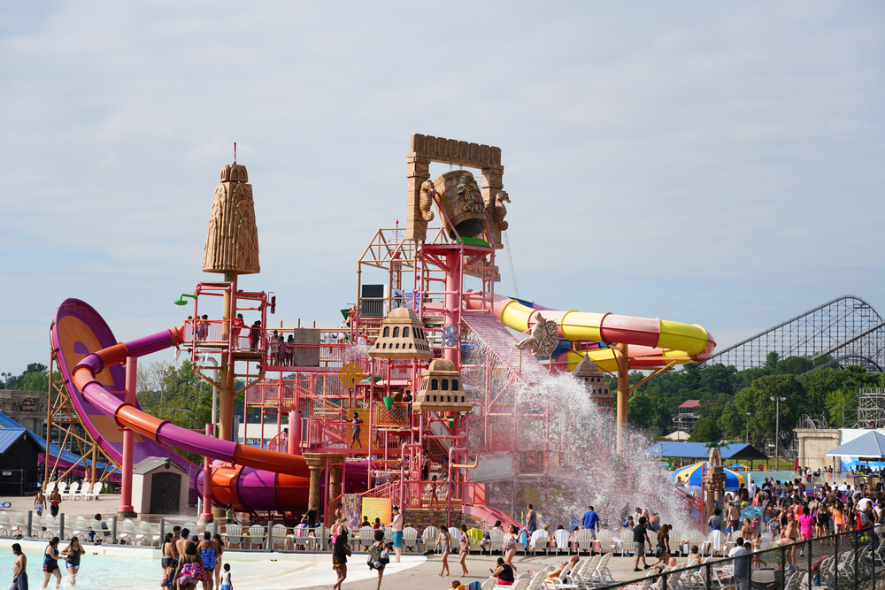 A large set of waterslides in different colors at a waterpark. There is also an area where you can climb up to the top of the slides, something splashing water on people, and a large bucket at the very top. There are people walking around the park, which is one of the best things to do in Wisconsin Dells. 