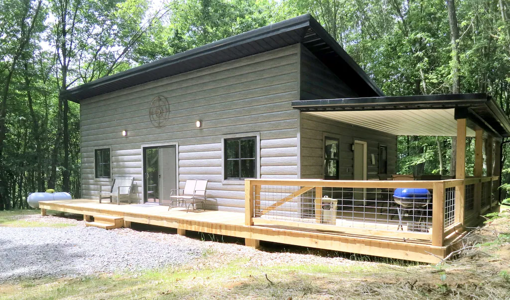 Grey Hocking Hills cabin with wooden wrap around decking, gravel parking lot in front. Bright blue barbecue grill on deck.