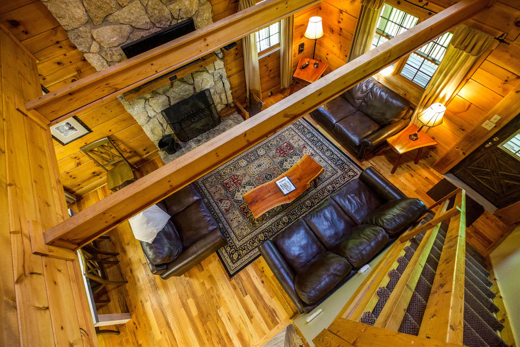 Overhead view of cabin living room, wooden walls, wooden floor, 2 leather couches, wood coffee table. Wooden stairs on right and stone fierplace on left.