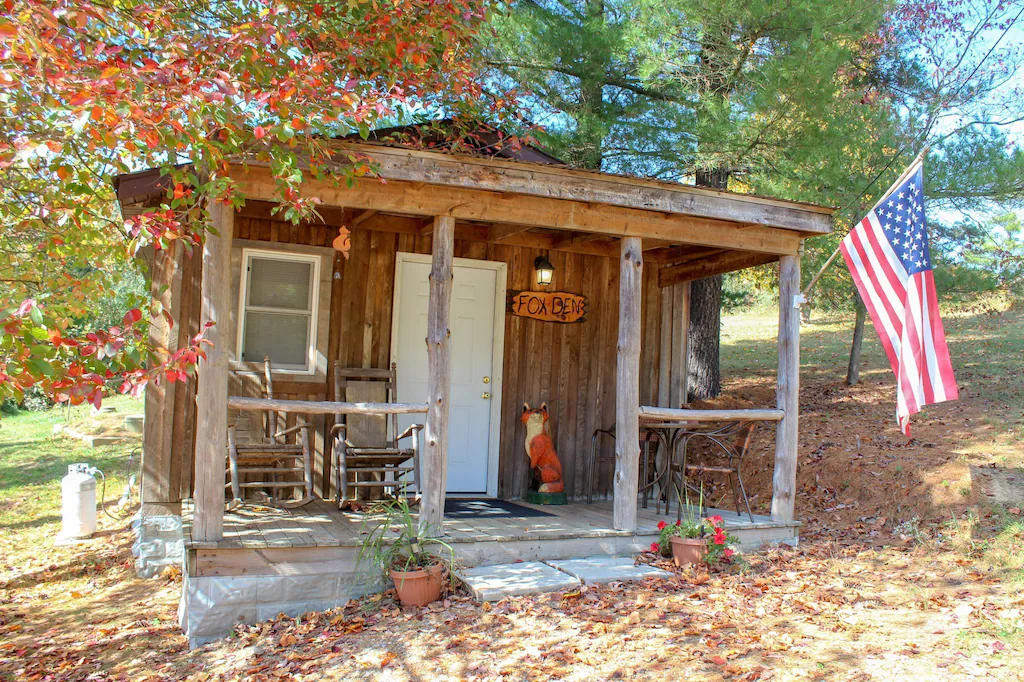Cute wood cabin with log columns, front porch with rocking chairs. Fox statue near front door, American flag flying on right.