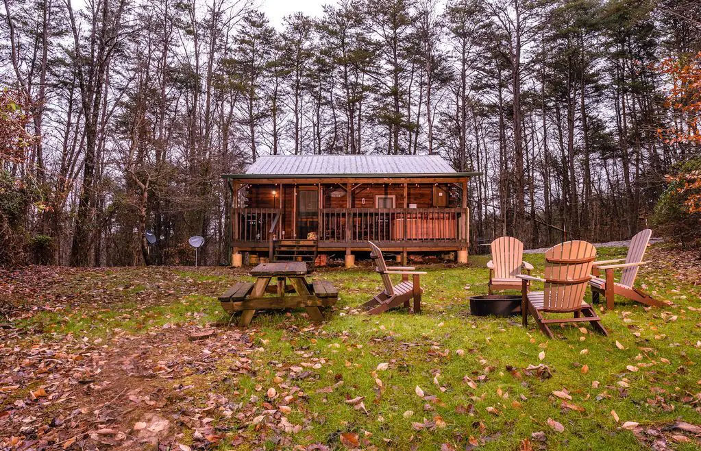 Log cabin with metal roof, large front porch with wood railing, winter trees in background, firepit and chairs and picnic table in front. Cabins in Hocking Hills.