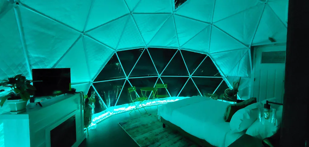Interior of geodesic dome living space with bed on right, kitchenette on left illuminated with bright blue hue.