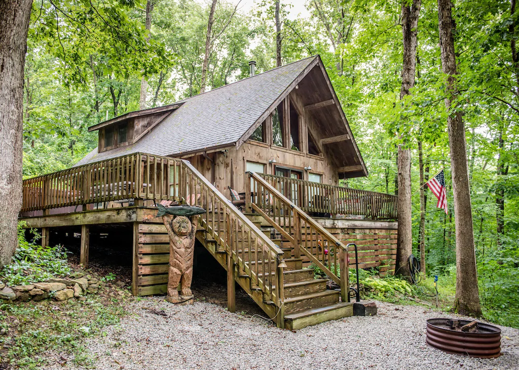 Log cabin in Ohio  with A-frame roof, large windows, balcony. Wooden statue in front next to stairs up to balcony.