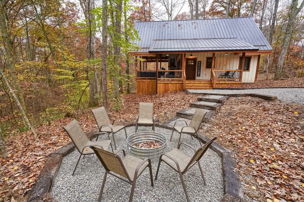 Cozy cabin in Ohio with brown metal roof and trees in background. Firepit and chairs in foreground.
