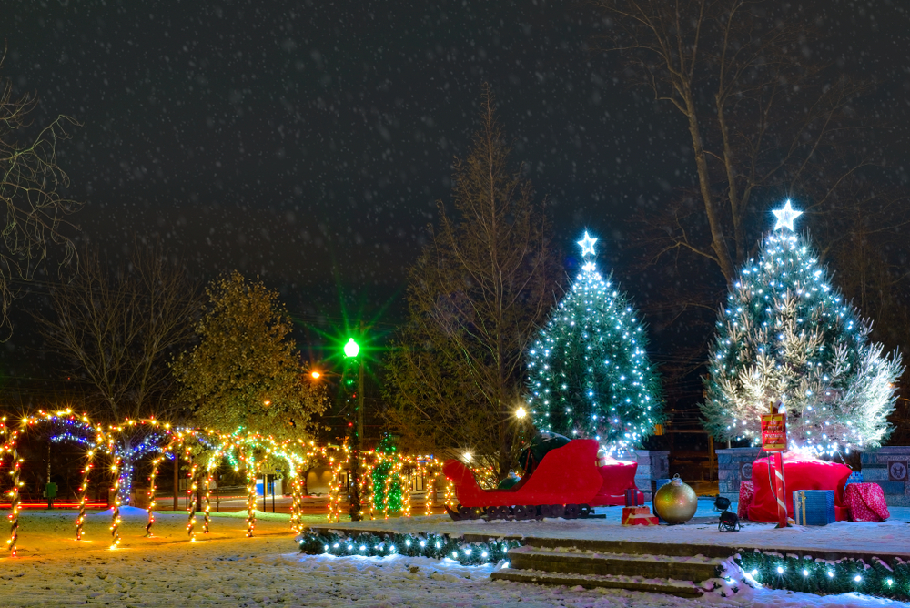 Two large Christmas trees with white lights, red sleigh and large ornaments in front of them, and yellow illuminated tree tunnel to the left.