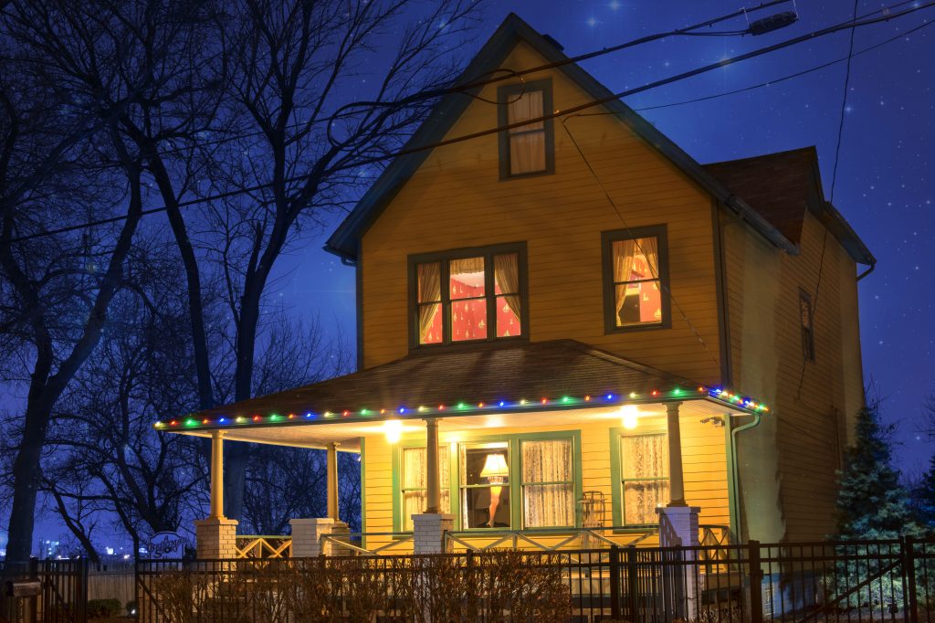 Yellow house with green trim, large front porch with multicolored lights, two windows on second floor illuminated. Christmas in Ohio.