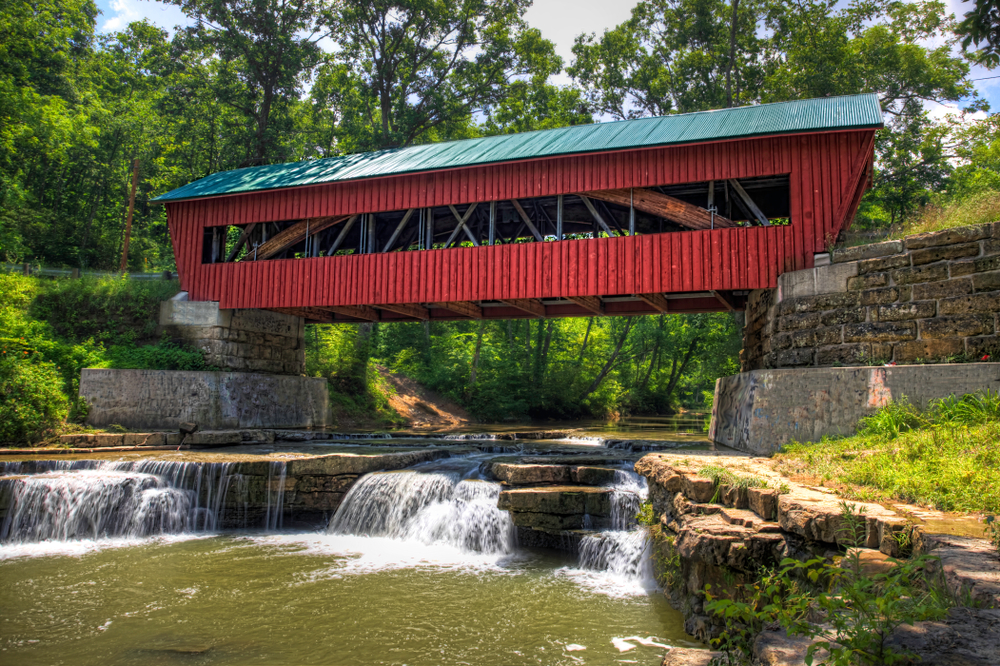 Red wooden bridge covered with green roof, spanning river with small waterfalls in foreground. Weekend getaways in Ohio.