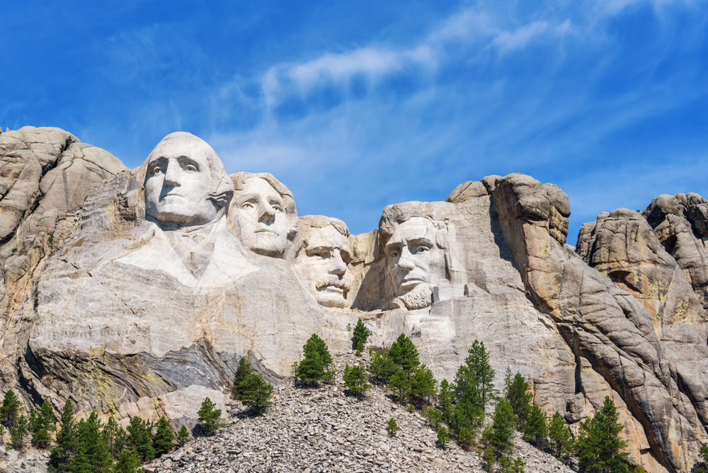 Four faces/heads of men carved into side of mountain with blue sky in background and evergreen trees at base of mountain.