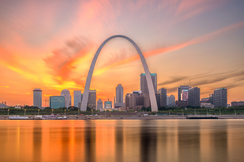 Orange sunrise sky with large cement arch monument and city skyscrapers in foreground, with calm waters and busy road in foreground.