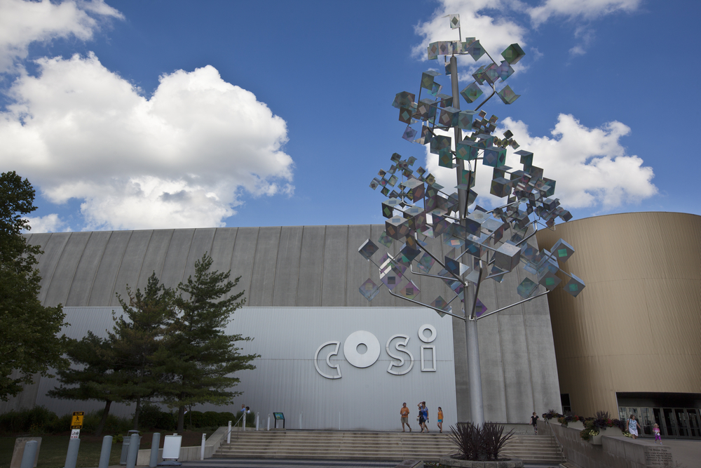 An industrial looking building, the Center of Science and Industry, is pictured as one of the best things to do in Columbus Ohio.  The building features the acronym COSI in silver lowercase letters.  A holographic statue that looks like a tree stands in front of the building.  