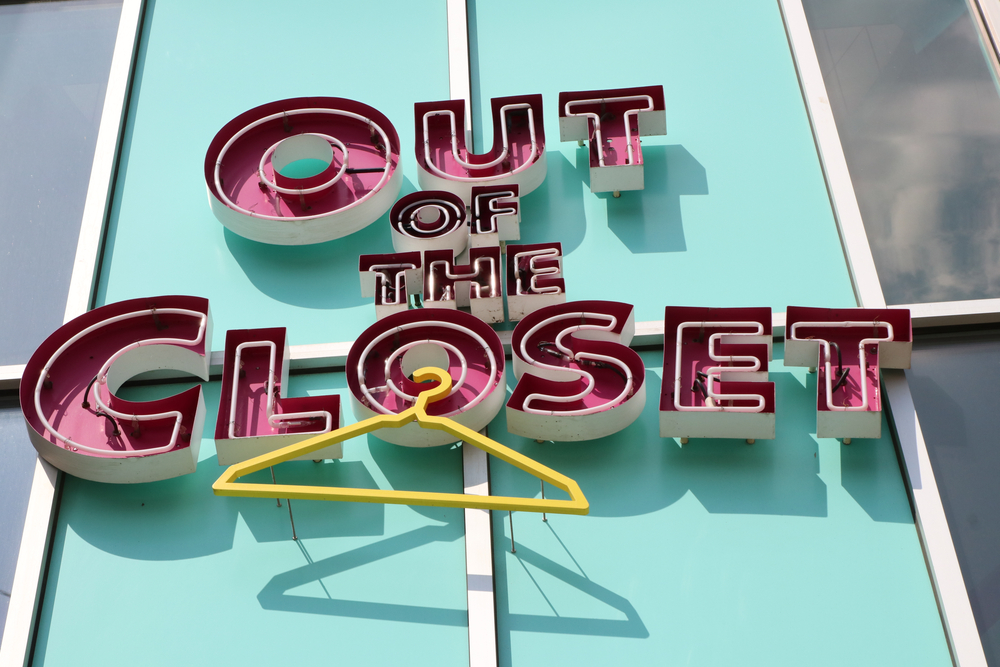 The sign for Out of the Closet Thrift Store is depicted.  It is teal with pink lettering and a yellow hanger hanging from the “O” in the word closet. 