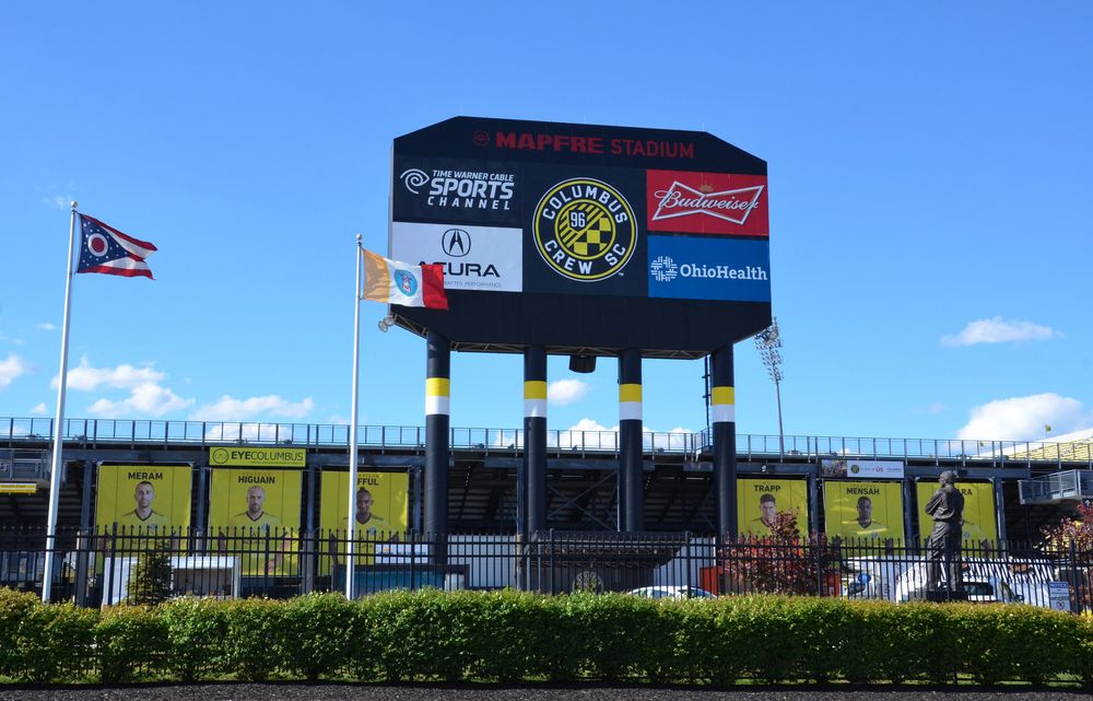 The Columbus Crew stadium is pictured with flags in front.  The players are pictured on banners, and the Crew’s sponsors are on a billboard.  Columbus Crew games are one of the best things to do in Columbus Ohio