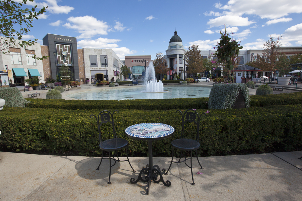 This photo features a wide shot of Easton mall.  In the foreground, there is a table with two chairs.  The mid ground has a fountain surrounded by a hedge.  The background has large stores like Macy’s and Tiffany & Co.  Easton is one of the best things to do in Columbus Ohio. 