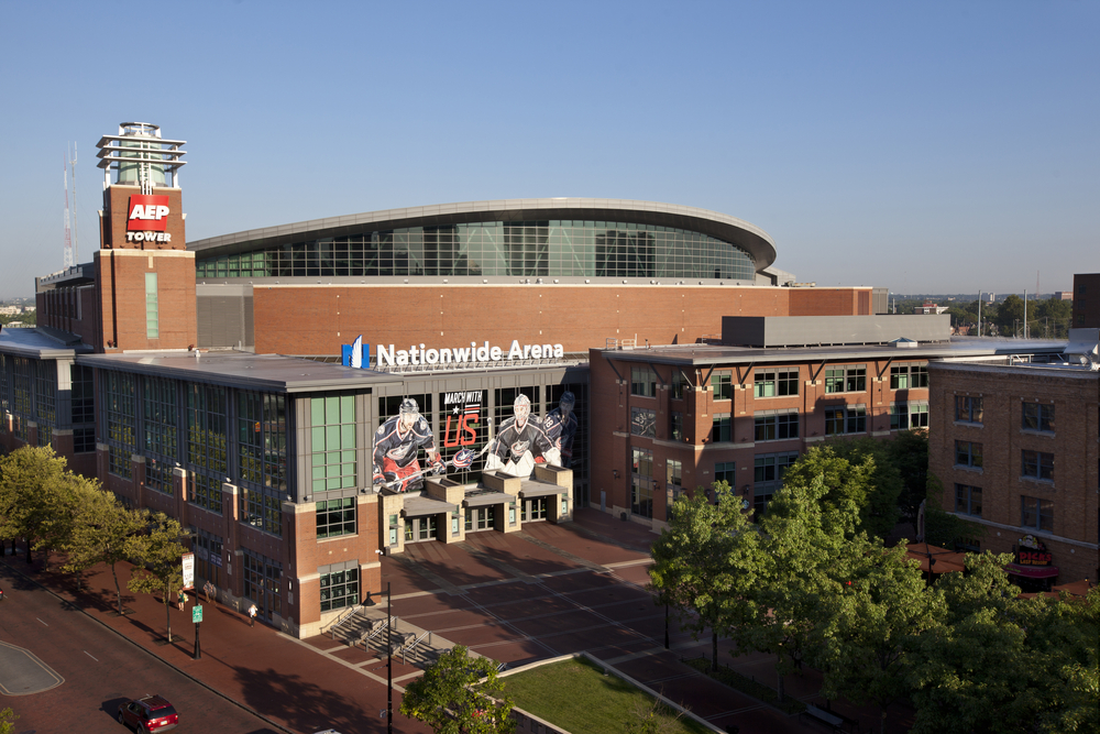 An aerial shot of Nationwide arena, one of the best things to do in Columbus Ohio, depicts the large, modern style building with hockey players depicted on the side. In front of the building is a courtyard with trees. 