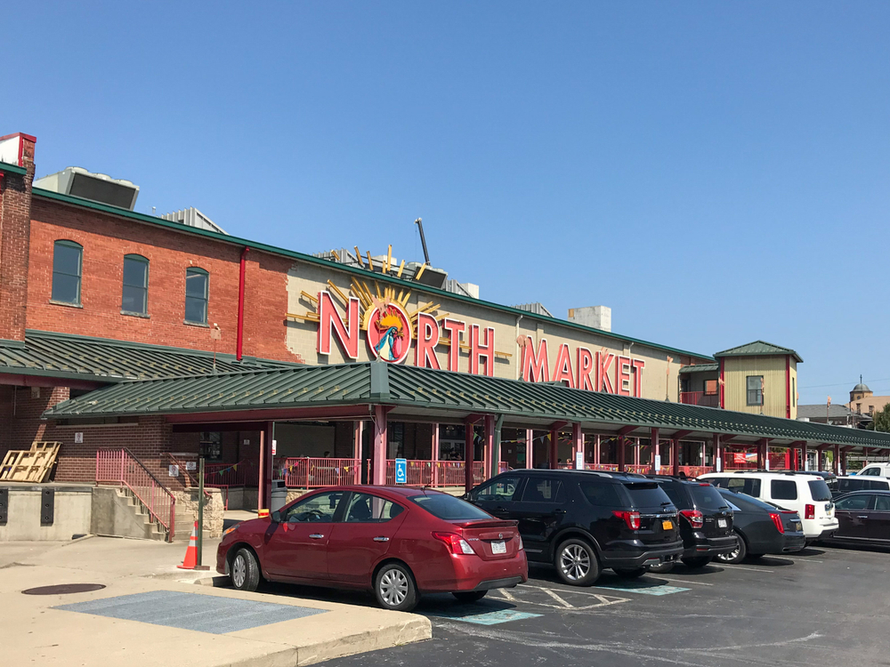 A building with green awnings is pictured with cars parked in front.  The building has the words “North Market” displayed in bold red lettering.  The “O” has a depiction of a rooster crowing inside.  