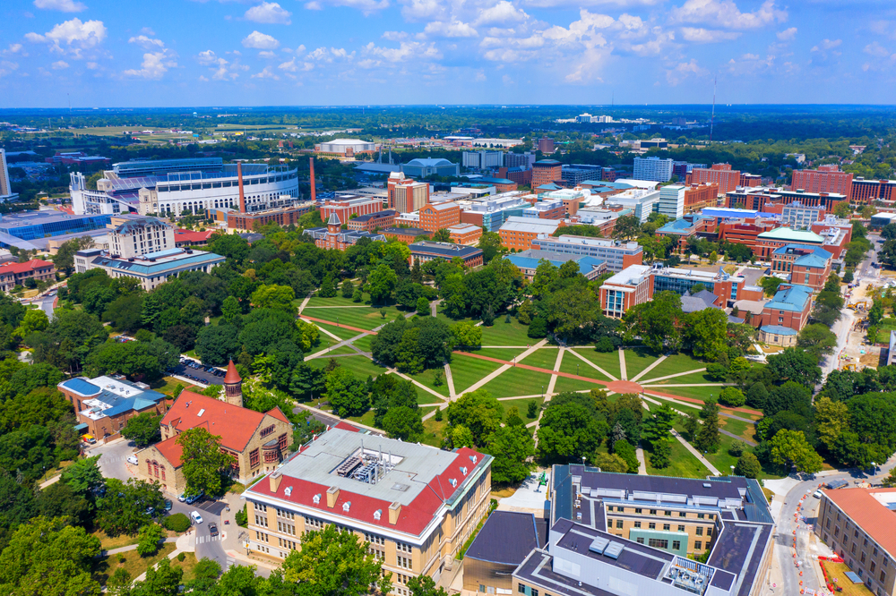 An aerial shot of The Ohio State University is depicted with many buildings and a lot of green space and trees.  The main quad on campus, the Oval, is the central focal point of the picture, notable for its abstract sidewalk pattern.  