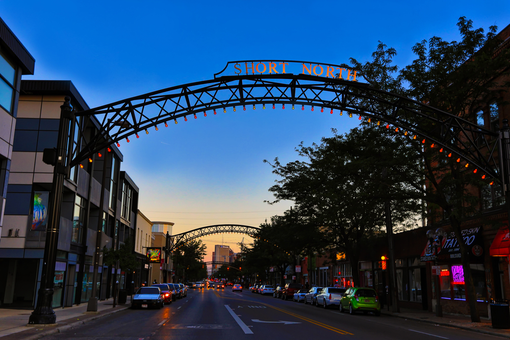 The short north, one of the best things to do in Columbus Ohio, is depicted at sunset.  Buildings line High Street as iron arches sprawl across the road with small lights on them.  