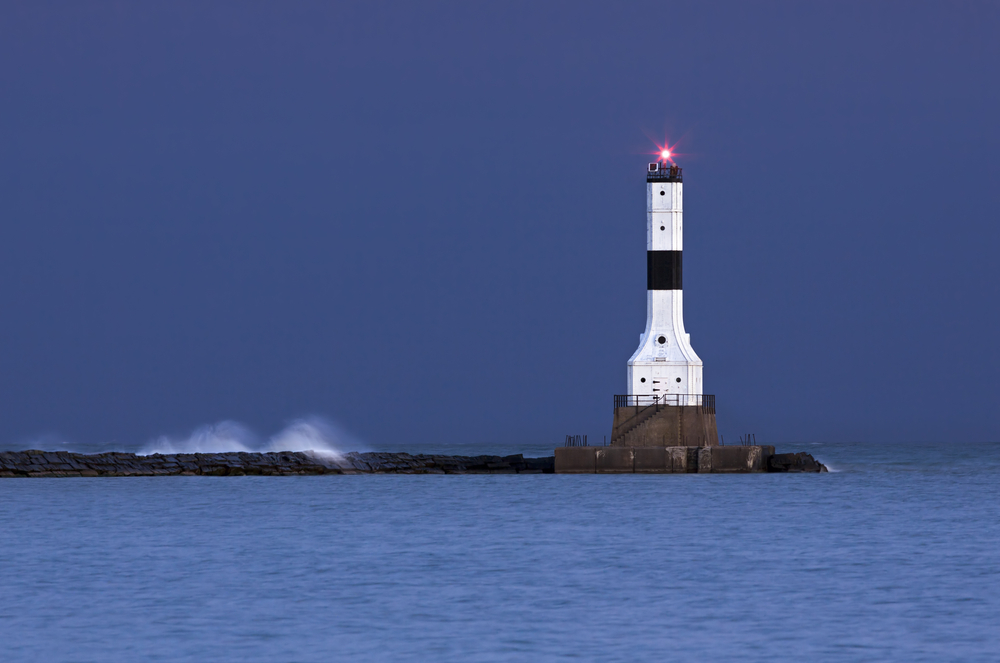 White and black lighthouse at end of stone wall with water splashing over it