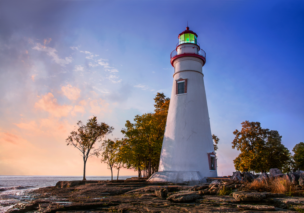 White lighthouse with red trim and light at top with trees surrounding it and water in background.