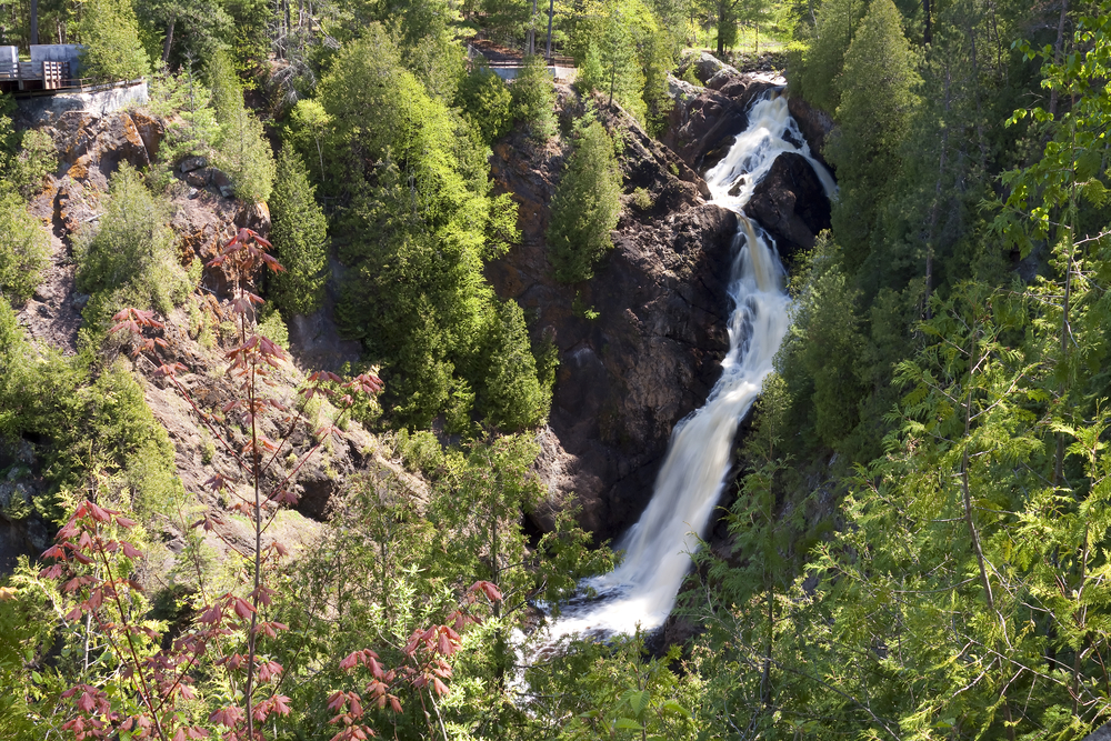 The Big Manitou Falls cascading over a tall rocky cliff side surrounded by tall trees with green leaves and other large rock formations. 