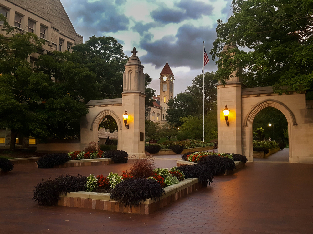 A brick courtyard with flower beds that have red and white flowers in them. There are also stone arches that lead to another part of the courtyard and trees. In the distance you can see a clock tower. One of the best weekend getaways in Indiana.