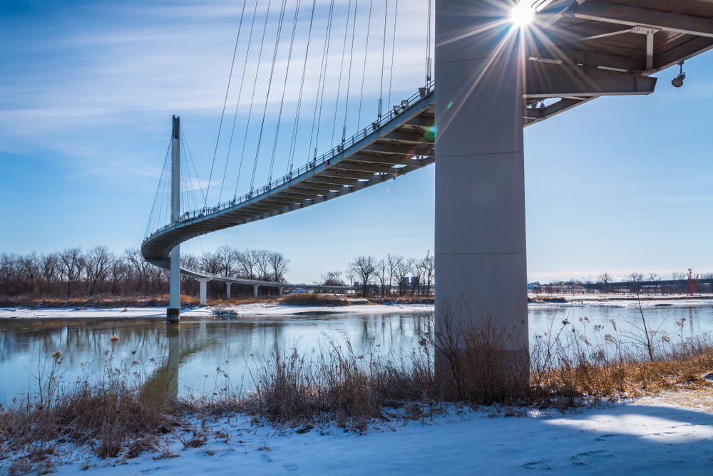 Looking at the Bob Kerrey Pedestrian bridge from the shores of the Missouri River. You can see the bridge curving over the river and going over a small island before continuing on. The bridge is white with suspension wires. The shore of the river is covered in snow. Its one of the most fun things to do in Omaha. 