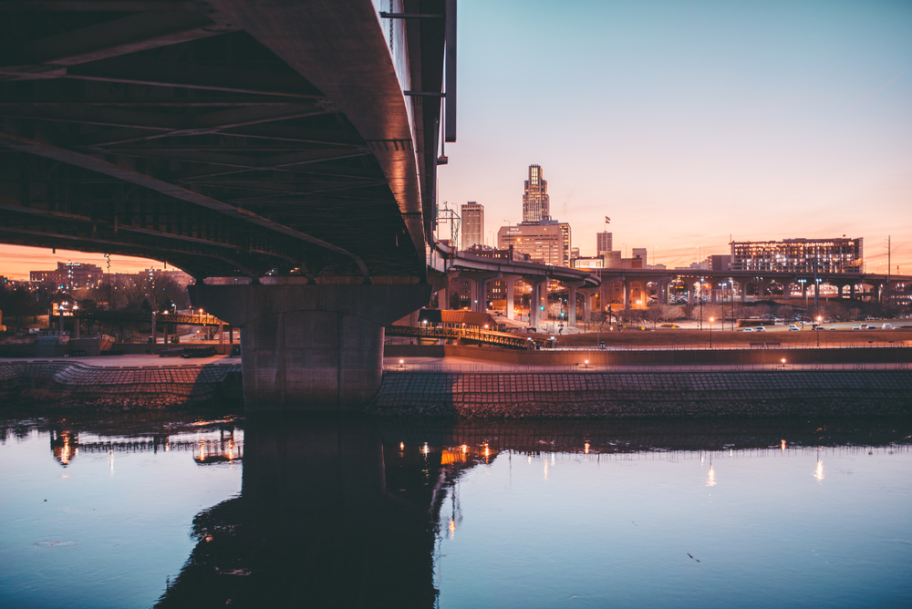 Looking at the skyline of Omaha from across the rive near the Bob Kerrey Pedestrian Bridge. It is twilight so the buildings are lit up and the bridge and river and dark. 