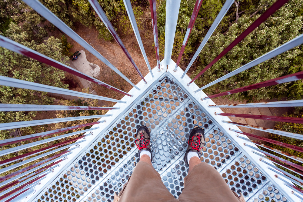 Looking down at someone's feet as they stand on a metal overlook tower. Below the tower you can see a dirt path and lots of trees. 