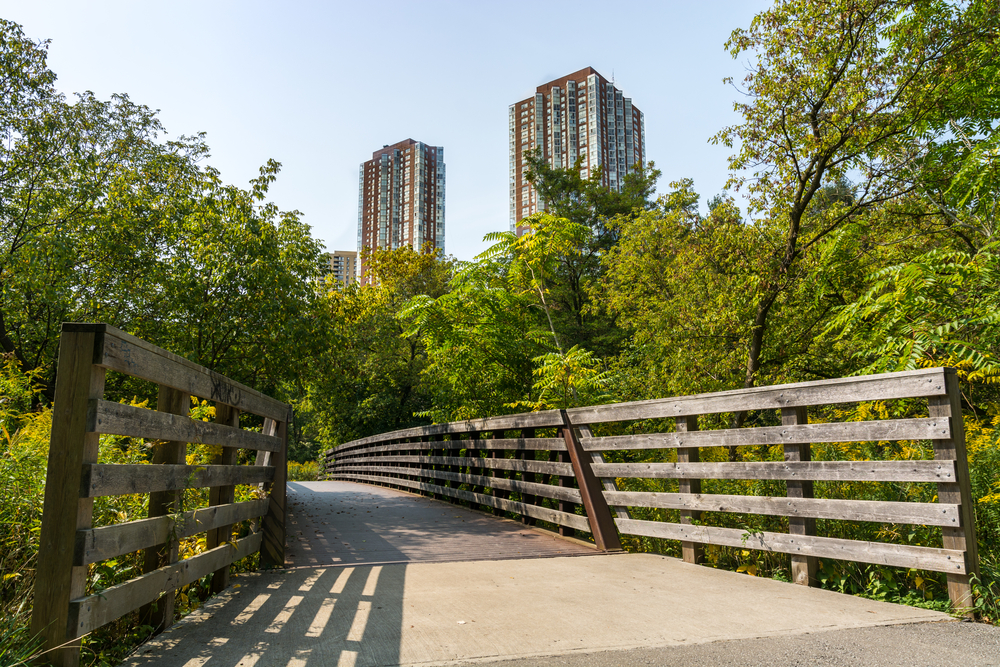 Looking down a path in the Fontenelle Forest, one of the fun things to do in Omaha. There is a path with wooden railings and all around it are trees. In the distance you can see two tall buildings. 