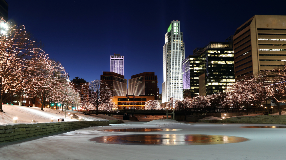 A view of the Omaha skyline from the Gene Leahy Mall, one of the fun things to do in Omaha. It is nighttime and all the buildings are lit up. There are twinkle lights in the trees and the river looks frozen over with snow on the ground. 