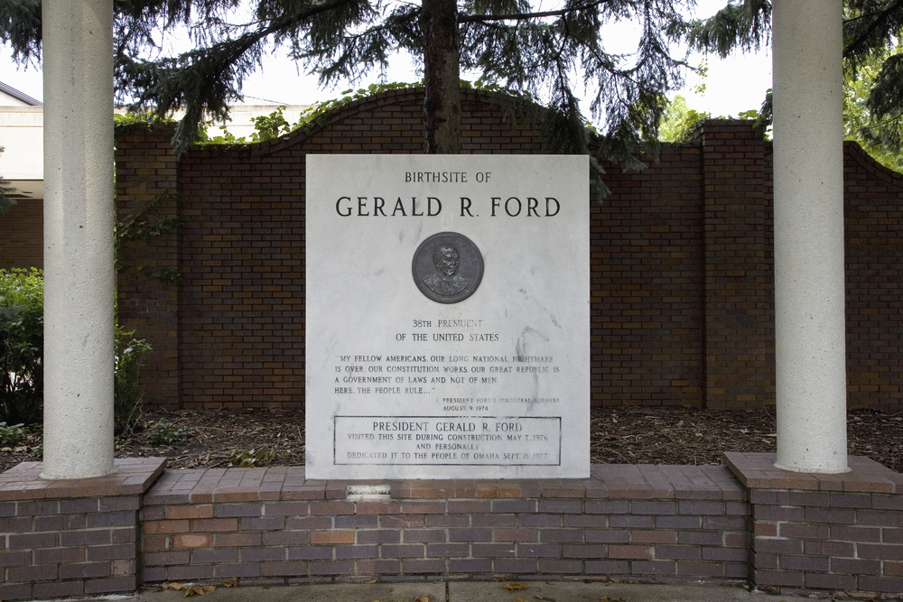 A plaque commemorating the birth site of President Gerald R. Ford. It is white marble and is sitting on a short dark brick wall with white columns. There is a metal coin with Fords face on it and the marble slab is engraved. 