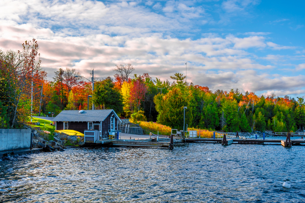 A small wooden cabin on the shores of a lake with a small dock. Behind the cabin you can see a dense forest with trees with green, yellow, orange, and red leaves. The sky is bright blue with thin clouds. One of the best Wisconsin road trips stops.