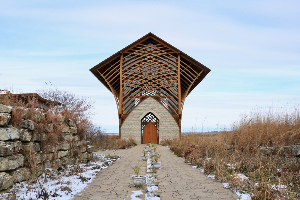 Looking at the Holy Family Shrine, one of the prettiest things to do in Omaha. It is a large shrine with stone, wood crisscrossing on the top, and a large roof. Around it there is a stone path, dead grass, and a stone wall. there are patches of snow on the ground. 