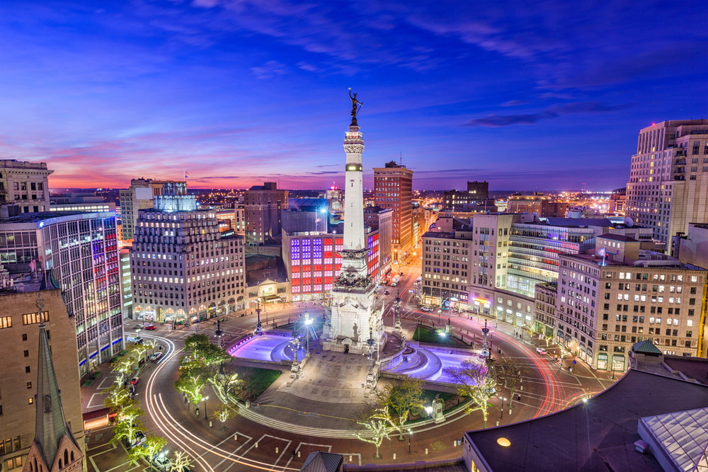 An aerial view of a large traffic square with a large statue in the middle of it. There are buildings all around the traffic square that are lit up and the sky is blue, purple, and pink because it is twilight.