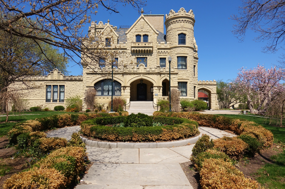 The front exterior of a large stone mansion that has a Scottish style. In front of it is a round pathway that has a shrub garden in the middle and shrubs all around the path. 