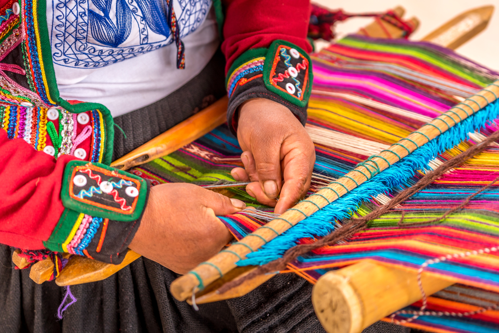 The hands of someone wearing a colorful Latin outfit while they weave using multicolored threads on a large loom. 