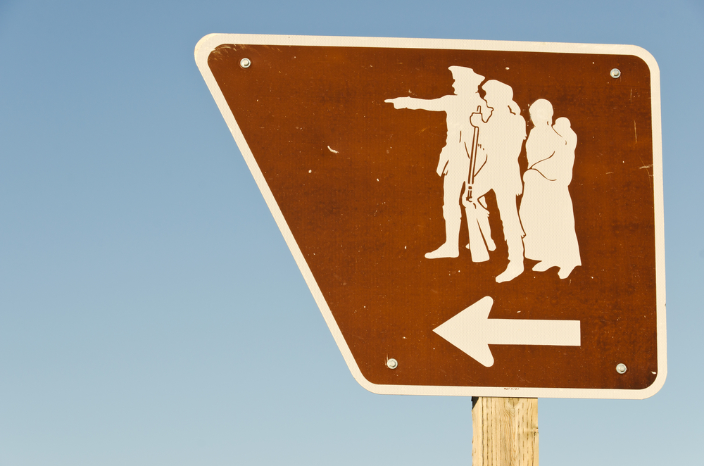 A brown sign on the side of the street. The sign has an odd shape and silhouettes of Lewis, Clark, and Sacagawea with an arrow pointing to the left. 